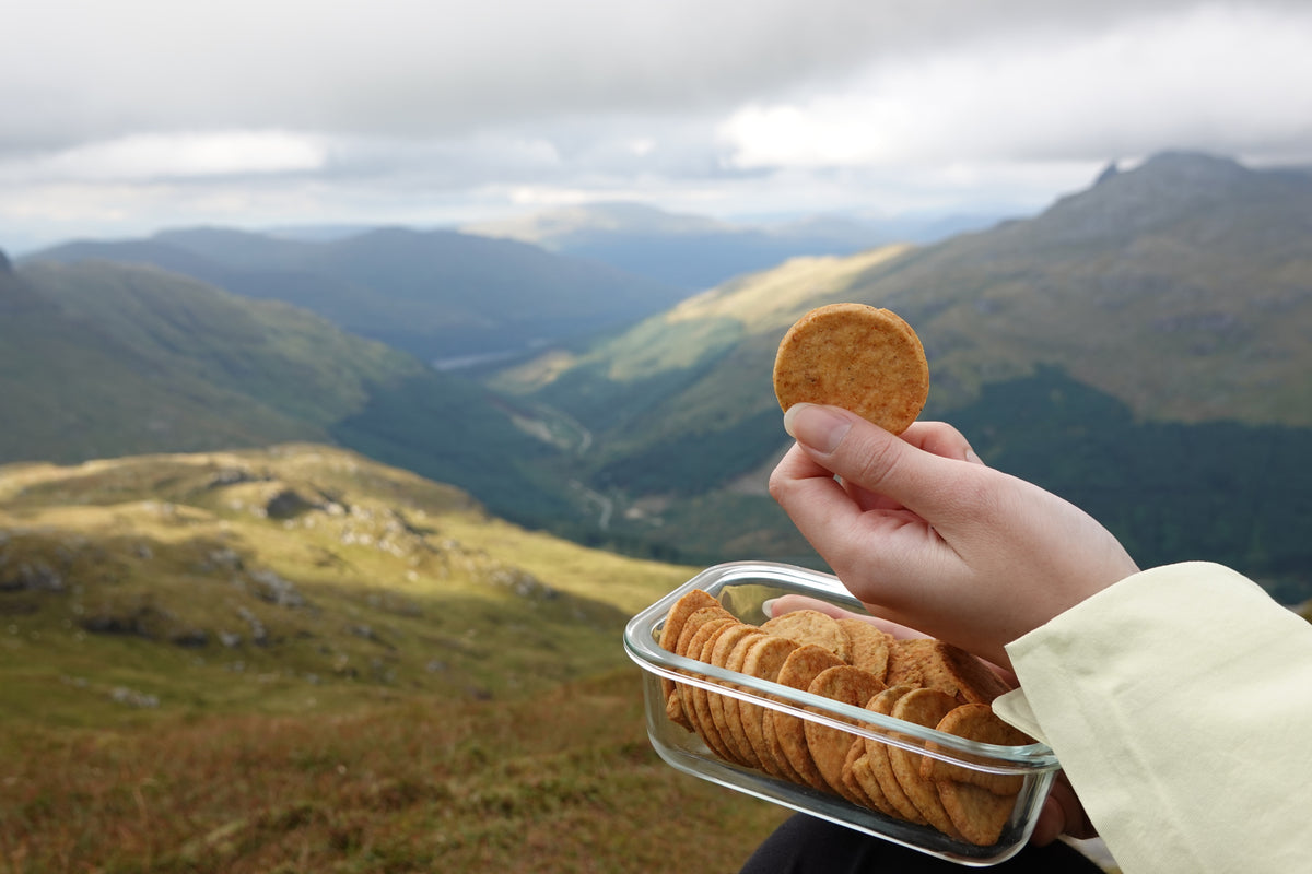 A walker in a pale yellow jacket sits on a hillside with a glass tub of TEMPTÉ tempeh snacks on their lap.